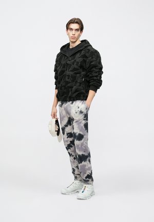 Felpa con zip in pile nero con motivo strutturato, abbinata a pantaloni della tuta tie-dye grigi e neri. Scarpe da ginnastica bianche e cappello beige in mano.