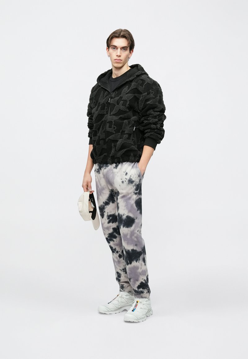 Mustast fliisist tõmblukuga kapuutsiga pusa tekstuurse mustriga, sobitatud hallide ja mustade lipsuvärvitud dressipükstega. Käes valged tossud ja beež nokkmüts.