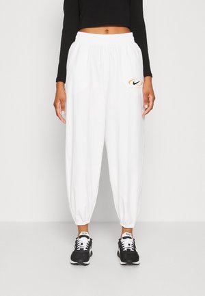 Personne portant un jogger blanc avec taille et chevilles élastiques, un petit logo Nike sur la cuisse gauche, un crop top noir à manches longues et des baskets noires et blanches.