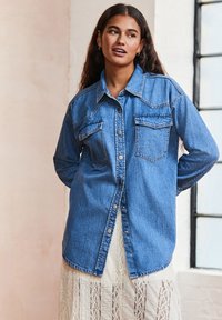 Camicia in denim blu con bottoni a scatto, maniche lunghe, due tasche sul petto, design strutturato e orlo grezzo, indossata sopra un vestito di pizzo.