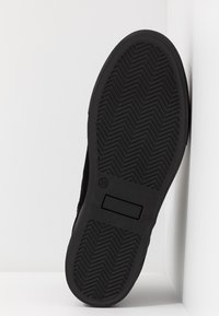 Semelle d'une chaussure noire avec un motif en chevrons, taille 38 indiquée, positionnée à la verticale contre un fond blanc en angle.