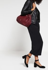 Czerwona skórzana torebka z marszczonym wzorem, z srebrnym łańcuszkiem na uchwycie, noszona z czarną skórzaną kurtką i czarnymi spodniami w kroju 3/4, zestawiona z czarnymi zamszowymi szpilkami.