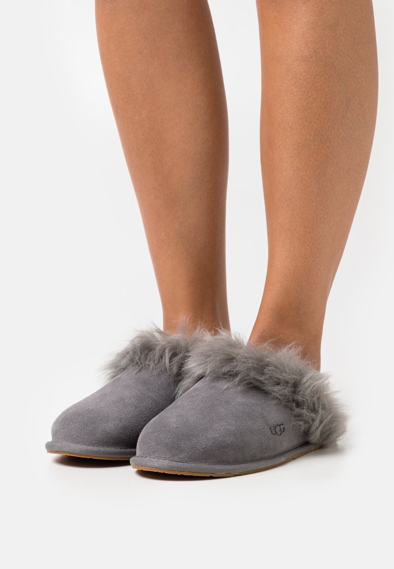 Pieds chaussés de chaussons slip-on en suède gris avec une bordure en fourrure douce et le logo UGG sur le côté, sur un fond blanc uni.