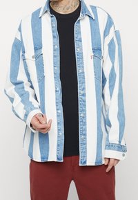 Camisa vaquera con rayas verticales alternas en azul claro y blanco. Cuenta con cierre de botones, bolsillos en el pecho y cuello y puños de contraste.