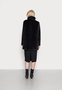 Sort faux fur frakke med høj krave, boxy snit og en kant, der falder ved midten af låret. Sammen med en knælang sort nederdel og ankelstøvler.