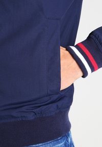 Handen i fickan på en marinblå jacka med ribbat avslut som har vita och röda ränder, bärs tillsammans med blå jeans.