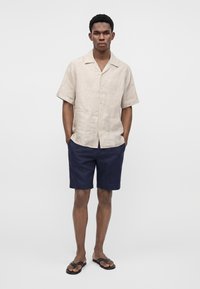 Homme portant une chemise beige à manches courtes boutonnée, un short bleu marine et des tongs noires, debout les mains dans les poches sur un fond uni.