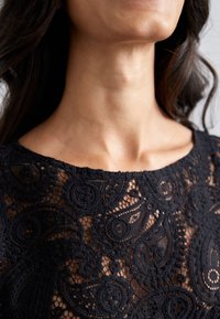Haut en dentelle noire avec des motifs paisley et floraux complexes, présentant une finition douce et texturée ainsi qu'un col rond.