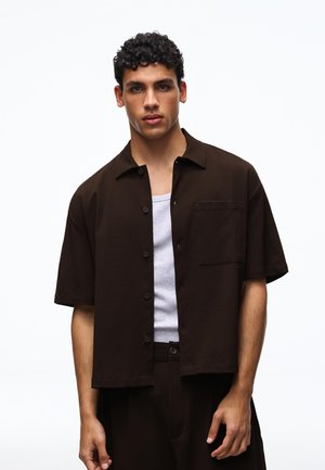 Młody mężczyzna o kręconych włosach, ubraną ciemnobrązową koszulę z krótkim rękawem zapinaną na guziki, na białym podkoszulku, stojący na jednolitym tle.