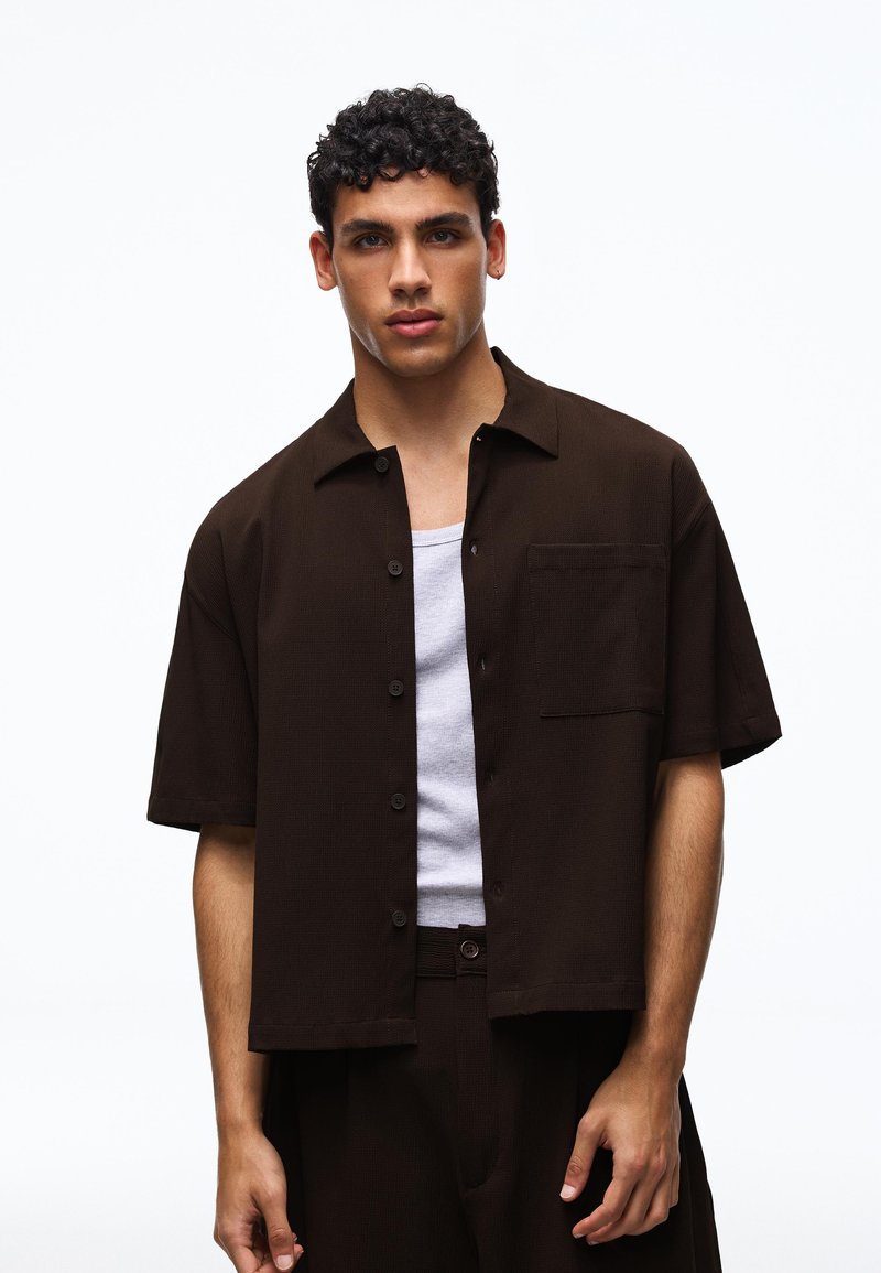 Jeune homme aux cheveux bouclés portant une chemise marron foncé à manches courtes boutonnée par-dessus un tee-shirt blanc, debout devant un fond uni.