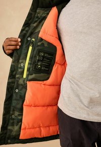 Tarnpufferjacke mit orangefarbenem Innenfutter, Reißverschlusstasche und wasserdichter Etikette. Hergestellt aus einem glatten, strapazierfähigen Stoff.