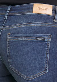 Blå denimjeans med en läderetikett i midjan, med framträdande sömmar och en liten svart varumärkesetikett på bakfickan.