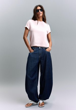 Frau trägt übergroße dunkelblaue Jeans, hellrosa hochgekrempeltes T-Shirt, schwarze Sonnenbrille und weiße offene High Heels, steht mit den Händen in den Taschen.