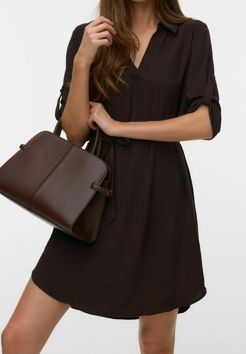 Femme portant une robe marron foncé tenant un sac à main en cuir marron foncé assorti avec des détails de manches retroussées.