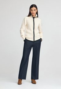 Crème kabelgebreide cardigan met een zwarte rits en groene accenten, gecombineerd met marineblauwe wijde pijpen broek en bruine enkellaarsjes.