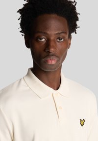 Polo shirt in crèmekleurige ribstof, met een gele geborduurde logo op de linkerborst, voorzien van een traditionele kraag en knopenlijst.