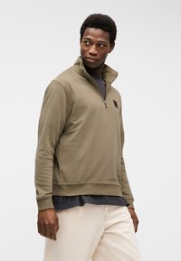 Junger Mann in beigefarbenem Sweatshirt mit Viertelreißverschluss über einem dunklen Hemd und heller Hose, schaut nach links vor einem einfarbigen weißen Hintergrund.