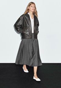 Giubbotto bomber in pelle nera con due tasche anteriori, blusa bianca, gonna plissettata grigia e ballerine bianche, su uno sfondo neutro.