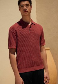 Polo shirt roșu cu mânecă scurtă și dungi, cu guler și placket cu trei nasturi, asortat cu pantaloni negri. Textura pare moale și țesută.