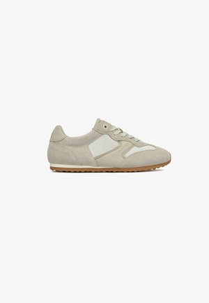 Zapatilla de perfil bajo en beige y blanco con paneles de ante y malla, cierre de cordones y suela de goma, vista lateral sobre fondo blanco.