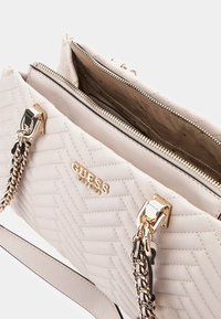 Borsa a mano color beige chiaro con texture a zig-zag, accessori in argento e tracolla a doppia catena. Presenta una chiusura a zip e un logo del marchio sul davanti.