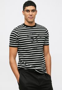 Vyras, vilkintis juodai baltą dryžuotą marškinėlį su užrašu "Karl Lagerfeld 21 Rue St-Guillaume Paris", juodas kelnes, rankas kišenėse.