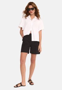 Biała koszula z krótkim rękawem, czarne strukturalne szorty, brązowe sandały ze złotymi detalami i czarne okulary przeciwsłoneczne. Prosty, casualowy letni strój.