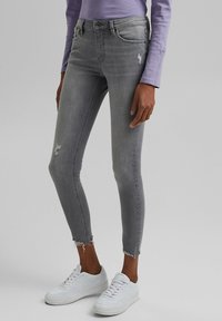 Jeans ajustados grises con detalles descoloridos, áreas desgastadas y dobladillos deshilachados. Combinados con zapatillas blancas y una camiseta de manga larga color púrpura claro.