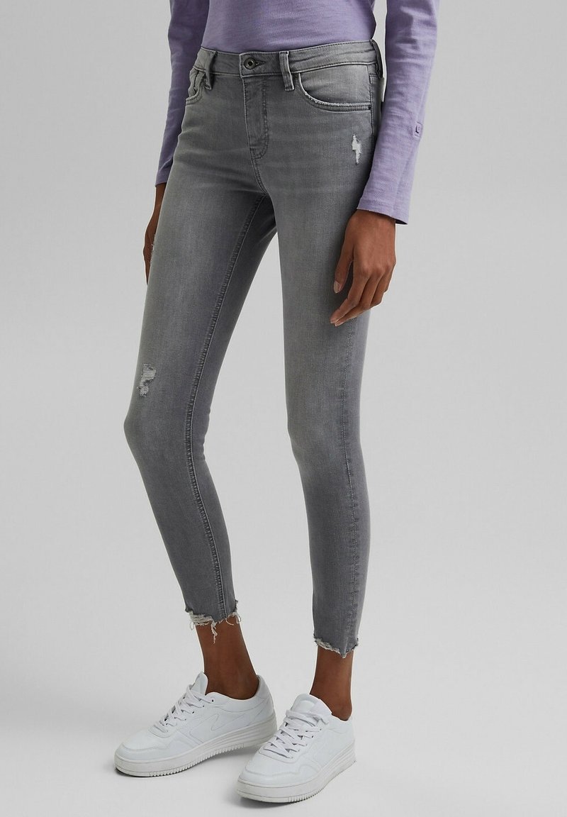 Jeans ajustados grises con detalles descoloridos, áreas desgastadas y dobladillos deshilachados. Combinados con zapatillas blancas y una camiseta de manga larga color púrpura claro.