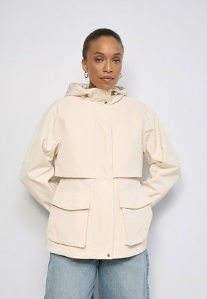 Mujer con chaqueta con capucha de color beige claro con grandes bolsillos delanteros y vaqueros azules, de pie frente a un fondo claro y liso.