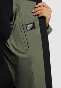 Groen olijfgroen jack met een lichte, gladde textuur aan de binnenkant. Voorzien van een zwart stoffen label met witte tekst en een contrasterende zwarte buitenlaag.