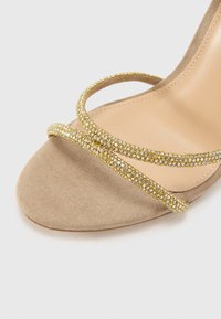 Svītrota augstpapēžu sandale no bežāda zamša, kas rotāta ar divām spirālveida siksnām, kuru apdari papildina dzeltenas un caurspīdīgas rhinestones.
