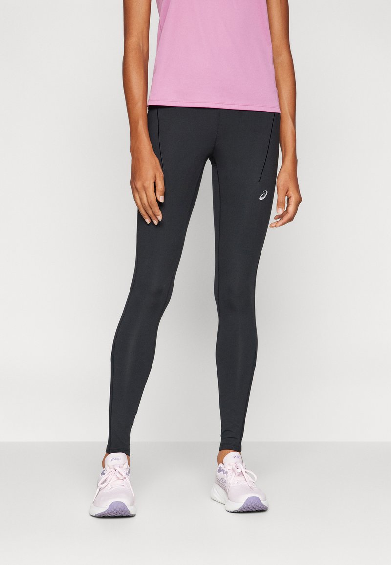 Zwarte legging gemaakt van een glad, rekbaar materiaal, met een aansluitende pasvorm en subtiele branding aan de zijkant, gecombineerd met lichtroze sportschoenen.