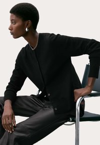 Černý kardigan se zapínáním na knoflíky a žebrovanými manžetami, nošený přes tmavý top, kombinovaný s elegantními černými koženými kalhotami, sedící na židli.