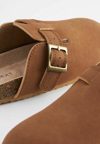 Brune ruskinds slip-on træsko med justerbar messing spænde og korksål, vist i nærbillede på en hvid baggrund.