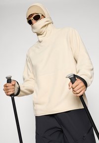 Cream fleece pullover with a high collar, black accent cuffs, and textured fabric. User holds trekking poles, wearing sunglasses and a beige beanie.