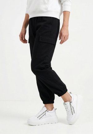 Person iført sorte cargo jogger bukser kombineret med hvide platformsneakers med sort tekst på snørebåndene.