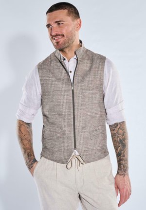 Mann mit kurzen Haaren und Tattoos auf beiden Armen, der eine beige Reißverschlussweste über einem weißen, hochgekrempelten Hemd und hellfarbene Hosen trägt.