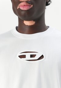 Camiseta blanca de algodón con un diseño de corte ovalado y detalles bordados. Textura suave; escote redondo.