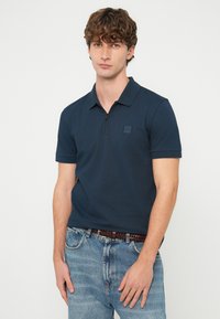 Νέος άνδρας με κατσαρά μαλλιά στέκεται φορώντας μπλε σκούρο πόλο με φερμουάρ, μέσα σε γαλάζια τζιν με καφέ πλεκτή ζώνη.