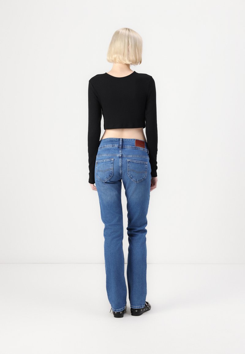 Schwarzes, langärmliges, cropped Oberteil, kombiniert mit mittel-hoch geschnittenen blauen Jeans, die über Gesäßtaschen und ein braunes Lederpatch am Bund verfügen.