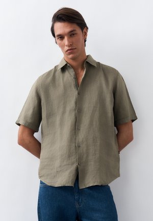 Νέος άνδρας με σκούρα μαλλιά, φορώντας πουκάμισο λαδί με κουμπιά και κοντά μανίκια και μπλε τζιν, στέκεται με τα χέρια πίσω από την πλάτη.