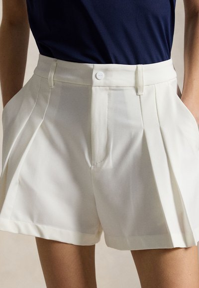 Beli kratki hlače z visokim pasom, krojeni, z gubami spredaj in zapiranjem z gumbi, nošeni s temno modro majico, roke v žepih, dolžina do sredine stegna.