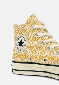 Žluté plátěné tenisky s vysokým střihem a bílou gumovou podrážkou, se špunty, šňůrkami a kulatou nášivkou s logem na boku.