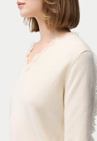 Cremefarbener Pullover mit V-Ausschnitt, verziert mit einem zarten Spitzenbesatz entlang des Halsausschnitts und einer weichen, glatten Textur. Lange Ärmel.