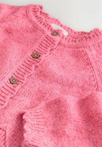 Różowy sweter z dzianiny, z okrągłym dekoltem, ribbowanym wykończeniem i drewnianymi guzikami z przodu. Tekstura wydaje się miękka i przytulna.