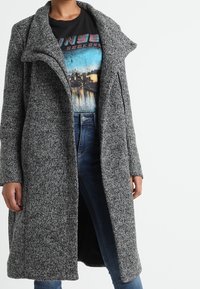 Cappotto lungo grigio in tessuto testurizzato, con un ampio collo, apertura frontale e tasche laterali, indossato sopra una maglietta grafica e jeans.