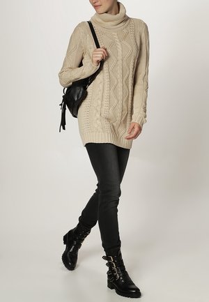 Maglione oversized beige a lavorazione di trecce con colletto alto, abbinato a jeans skinny neri e stivaletti neri con cinturini a fibbia.