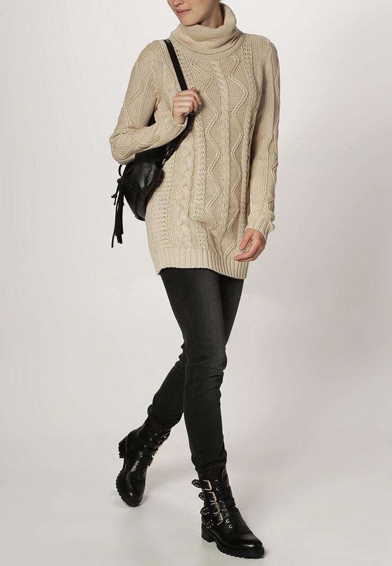 Beige kabelgebreide oversized trui met een col, gecombineerd met zwarte skinny jeans en zwarte enkellaarsjes met gespsluitingen.