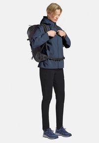 Marineblaue wasserdichte Jacke mit hohem Kragen, verstellbaren Riemen und einem Rucksack. Schwarze Slim-Fit-Hose und blaue Sportschuhe vervollständigen den Look.